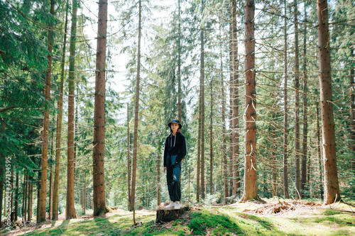 Woman hiker in panama and black casual clothes stands on a stump against the backdrop of a pristine forest with moss on ground, looking at camera with a serious face.Hipster girl hiking in the woods
