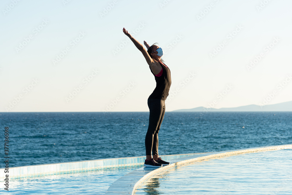 Obraz premium Young woman with protective surgical face mask performing yoga stretching exercises outdoor during covid-19 coronavirus pandemic
