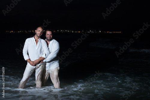 Gay couple of men happy and dressed in white bathe in the sea on the beach at night
