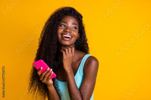 Photo portrait of african american woman with long wavy hair holding pink smartphone in two hands laughing wearing blue singlet isolated on vivid yellow colored background