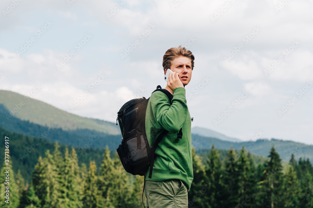 Serious young hipster in casual clothes and with a backpack talks on the phone in the mountains and looks sideways against the background of a mountain landscape with forest and hills.
