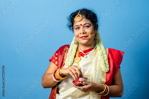 Married Indian Bengali brunette woman wearing traditional red and white sari is looking her face in a mirror while applying sindoor on her forehead.
