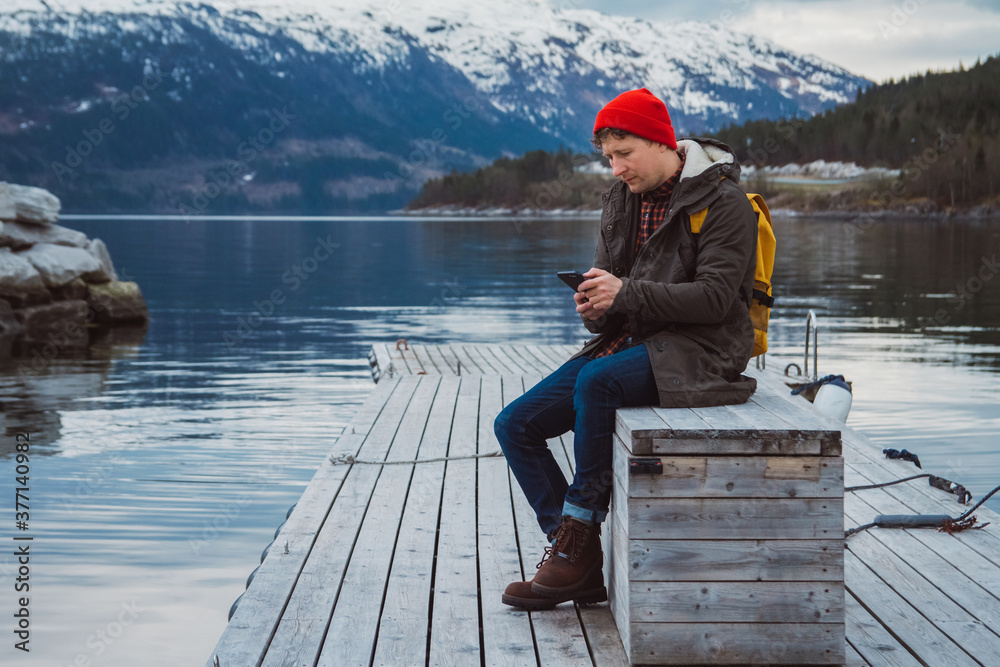 Young man looking into the smartphone with a yellow backpack wearing a red hat sitting on wooden pier on the background of mountain and lake Young man looking into the smartphone with a yellow backpack wearing a red hat sitting on wooden pier on the background of mountain and lake