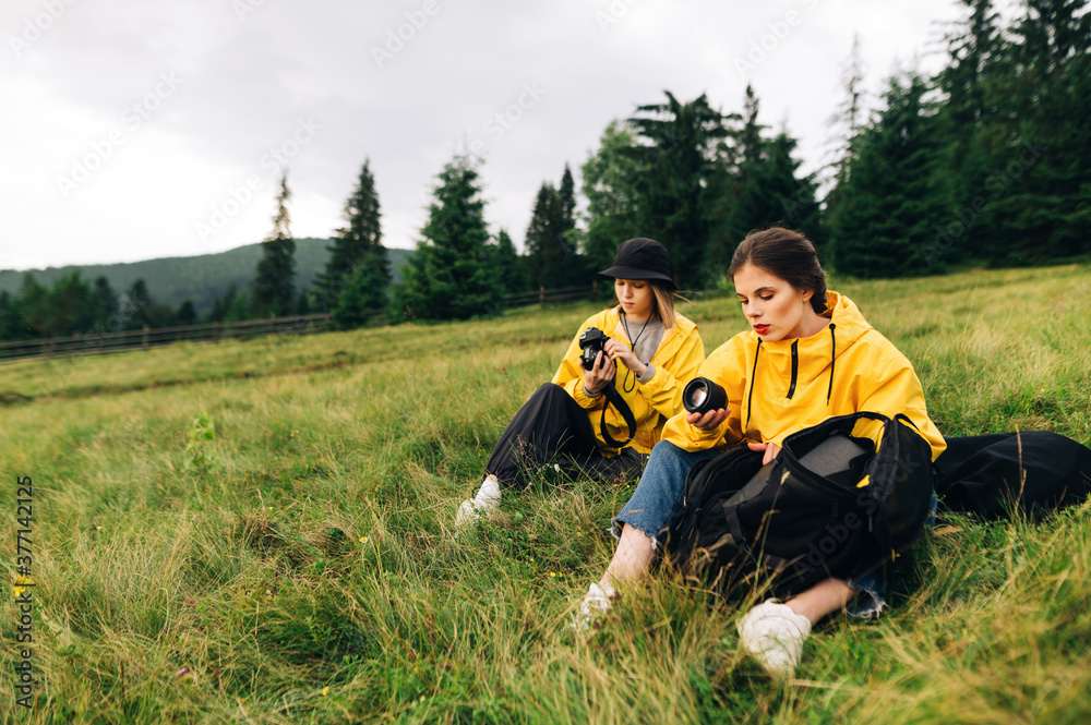 Two women photographers in yellow raincoats are sitting on a meadow in the mountains with photography equipment in their hands, one holding a lens and the other a camera.
