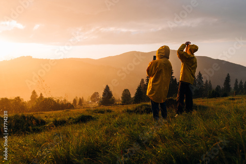 Two hikers in raincoats stand in the evening rain at sunset in the mountains and take photos of the unreal landscape on a smartphone. Hikers in the rain at sunset look at the beautiful landscape.