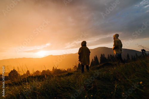 Two hikers stand in the rain on a meadow in the mountains and watch the incredible orange sunset. Two hikers in raincoats and sunset in the rain on a background of beautiful landscape.