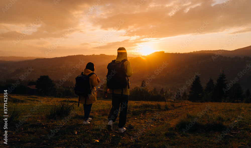 Two hikers in yellow raincoats descend from the mountain down on the background of a beautiful mountain landscape at sunset in the rain. Hikers go in rainy weather in the mountains at sunset