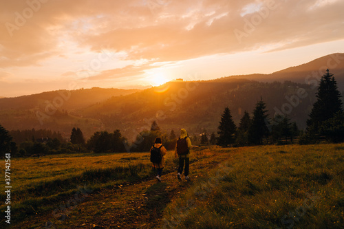 Two tourists in raincoats descend from the mountain in rainy weather against the backdrop of an unreal sunset. Hikers walk down the meadow on a background of sunset in the rain and mountain landscape