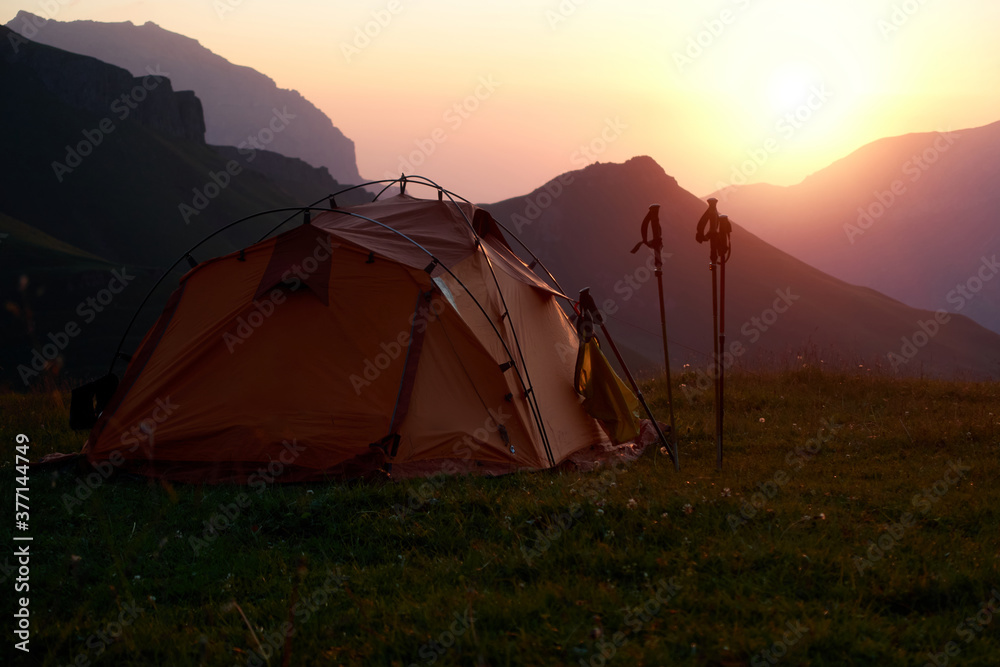 Tourist tent against the background of mountains at sunrise.
Tourist tent and trekking poles on the background of mountains at sunrise. Dawn in the mountains. Tourist camp in the mountains.