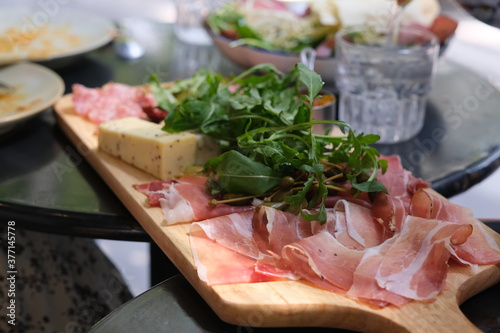 close up cured meats, arugula and cheese on wooden plate under sunlight. Traditional Italian food. Defocused background