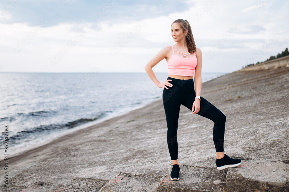 Slim positive girl. Attractive caucasian athletic girl in sportswear is standing near the sea during outdoors workout and smiling. Sporty and healthy life style Slim positive girl. Attractive caucasian athletic girl in sportswear is standing near the sea during outdoors workout and smiling. Sporty and healthy life style