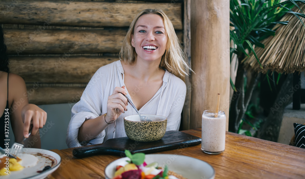 Portrait of cheerful blonde young woman looking at camera enjoying tasty food on breakfast keeping vegan diet, cropped image of hipster girl eating meal during lunch satisfied with dishes and drinks