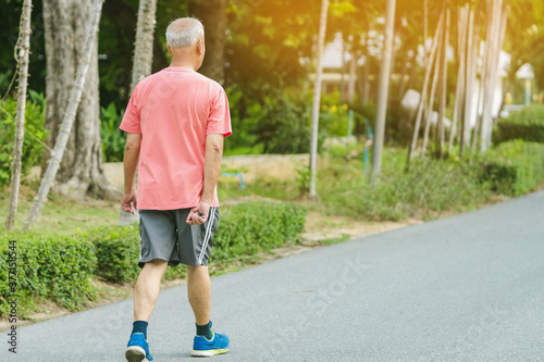 Back view portrait of a Asian elderly man in fitness wear walking and jogging for good health in public park. Senior jogger in nature. Older Man enjoying Peaceful nature. Healthcare concept.