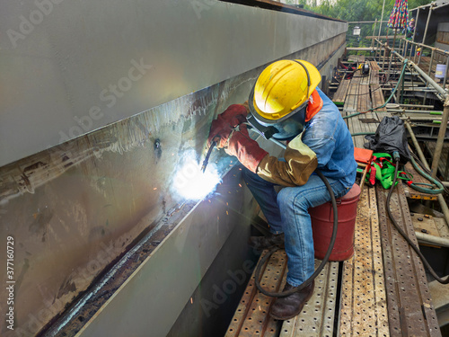 The welder is welding a steel structure work with process Flux Cored Arc Welding(FCAW) and dressed properly with personal protective equipment(PPE) for safety, at industrial factory.