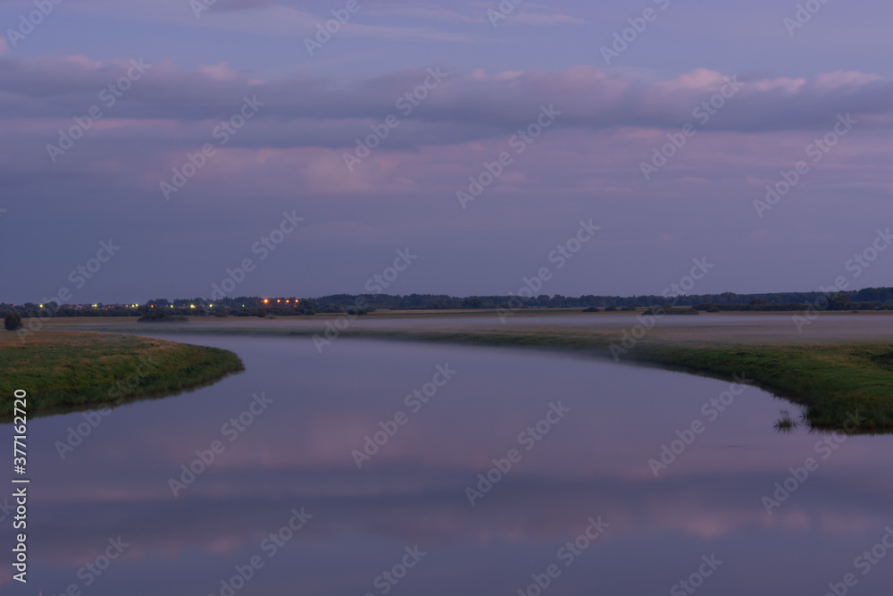 Summer evening landscape with a river in fog.River with green grassy foggy banks and reflection of clouds in the water with a forest and a village on the horizon Summer evening landscape with a river in fog.River with green grassy foggy banks and reflection of clouds in the water with a forest and a village on the horizon