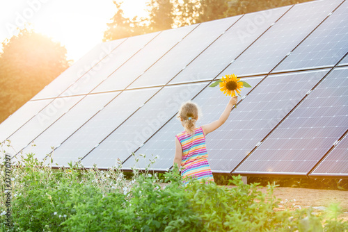 Young 6 year old blonde girl child standing in front of small solar panel farm in countryside. Renewable energy concept. Bright yellow warm sun lens flare.