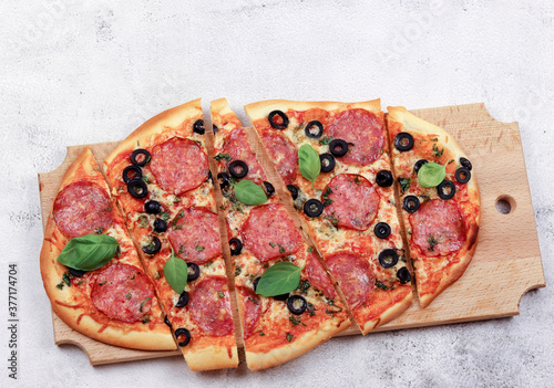 Sliced roman square pizza with cheese, sausage, olives and tomatoes on a round wooden cutting board on a light gray background. Top view, flat lay photo