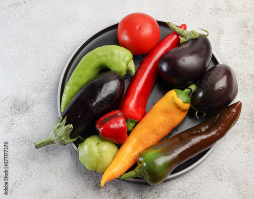 Assorted raw vegetables - bell peppers, tomatoes, hot peppers, eggplant on a round plate on a light gray background. Top view, flat lay