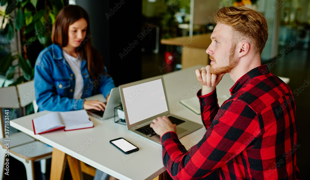 Pensive male programmer sitting at table desk with blurred female colleague pondering on update software system on mockup laptop computer with copyspace area for internet advertising, blank mobile Pensive male programmer sitting at table desk with blurred female colleague pondering on update software system on mockup laptop computer with copyspace area for internet advertising, blank mobile