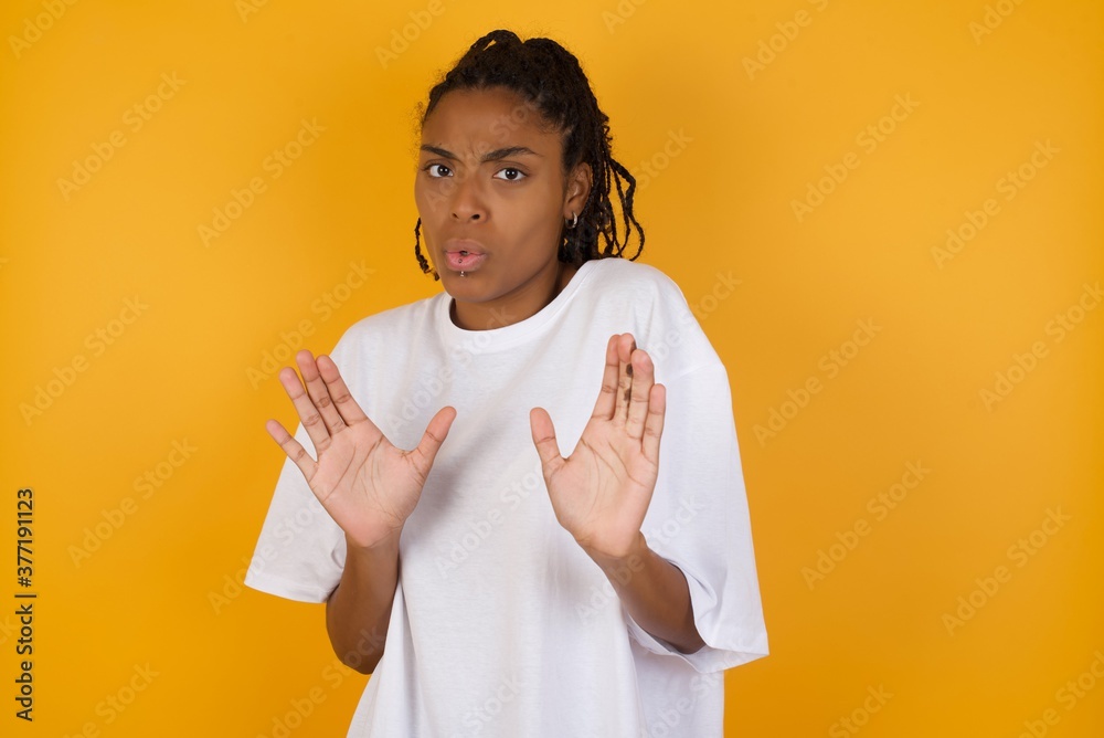 Young dark skinned woman with braids hair wearing white t-shirt over yellow background Moving away hands palms showing refusal and denial with afraid and disgusting expression. Stop and forbidden. Young dark skinned woman with braids hair wearing white t-shirt over yellow background Moving away hands palms showing refusal and denial with afraid and disgusting expression. Stop and forbidden.