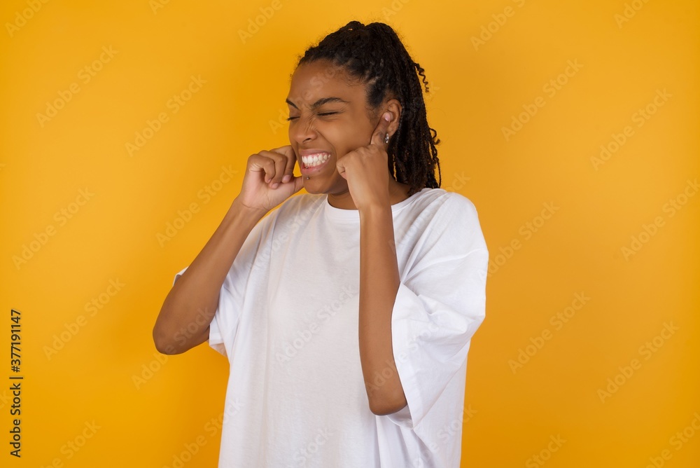 Young dark skinned woman with braids hair wearing white t-shirt over yellow background covering ears with fingers with annoyed expression for the noise of loud music. Deaf concept. Young dark skinned woman with braids hair wearing white t-shirt over yellow background covering ears with fingers with annoyed expression for the noise of loud music. Deaf concept.