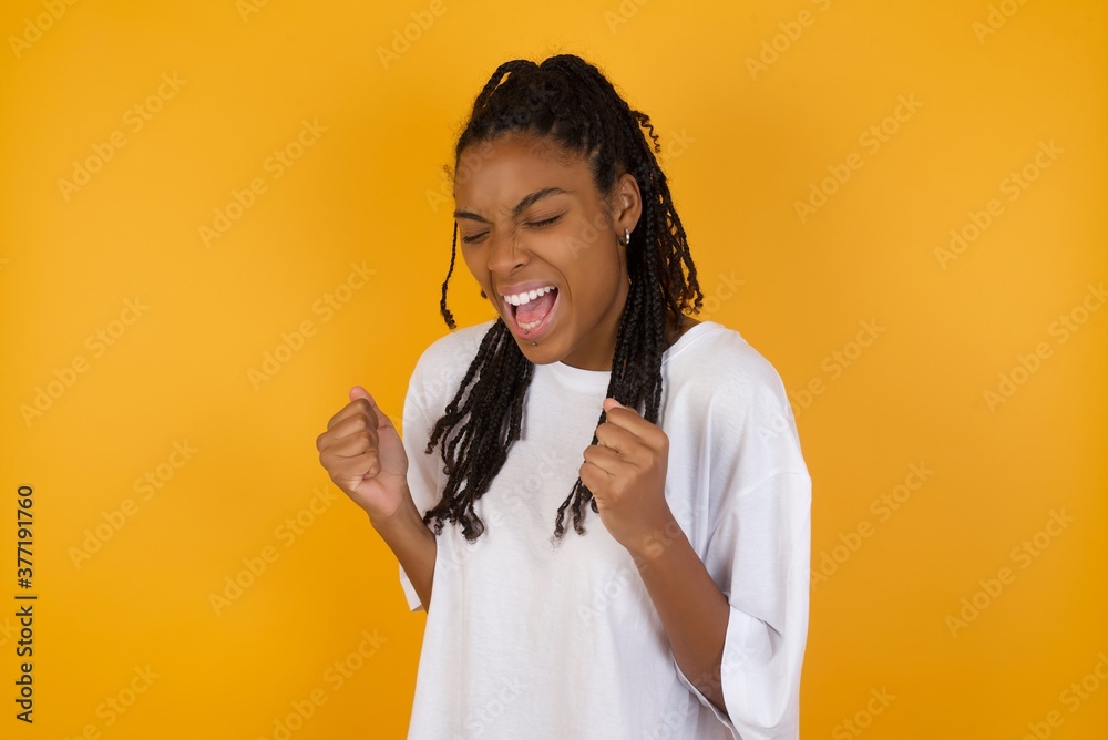 Young dark skinned woman with braids hair wearing white t-shirt over yellow wall very happy and excited doing winner gesture with arms raised, smiling and screaming for success. Celebration concept. Young dark skinned woman with braids hair wearing white t-shirt over yellow wall very happy and excited doing winner gesture with arms raised, smiling and screaming for success. Celebration concept.