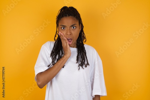 Shocked Young dark skinned woman with braids hair wearing white t-shirt over yellow wall looks with great surprisment being very stunned  astonished with unexpected news  . Facial expressions concept.