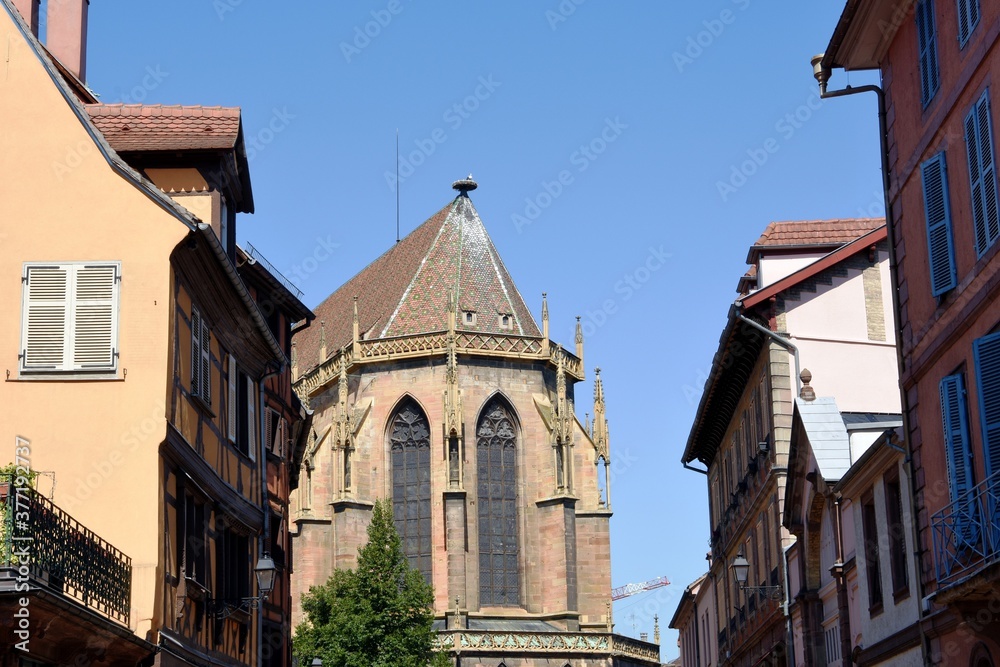 Narrow streets of medieval and early Renaissance buildings with window boxes draped with blooms in Colmar, town in the Grand Est region of northeastern France. On the background is St Martin's 