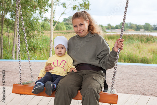 Two Caucasian girls of one year and twenty years old sitting together on swing, sisters play on playground