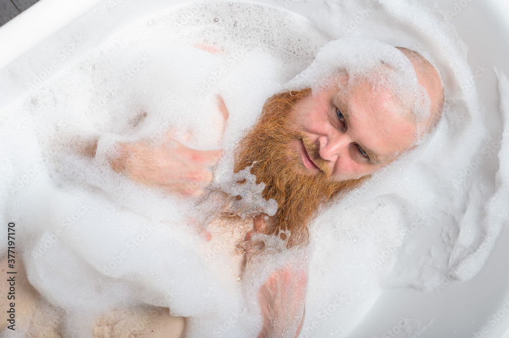 Top view of a bald man with a red beard splashing in the foam bath. Humorous photo. A parody of glamorous girls. Top view of a bald man with a red beard splashing in the foam bath. Humorous photo. A parody of glamorous girls.