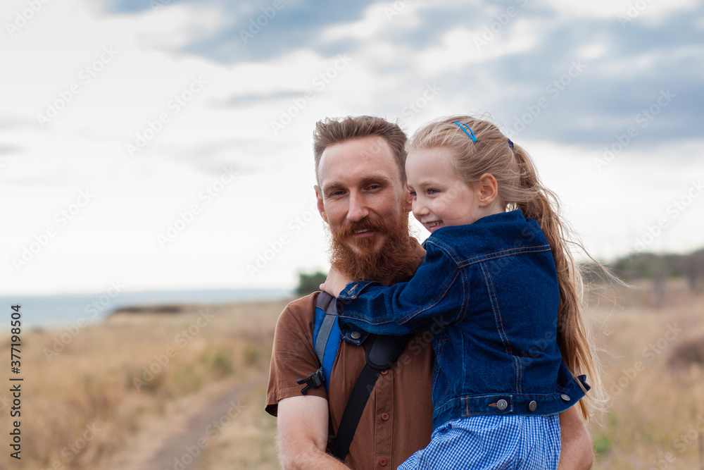 Father with daughter in hands standing on a hill with nature lanscape. Little blonde girl smiling and hugging with dad outdoor. Loving child embrace her daddy. Father's Day family greeting card. Father with daughter in hands standing on a hill with nature lanscape. Little blonde girl smiling and hugging with dad outdoor. Loving child embrace her daddy. Father's Day family greeting card.