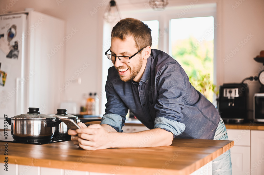 attractive nerd guy with glasses in stylish kitchen follows a recipe or reads news on the phone attractive nerd guy with glasses in stylish kitchen follows a recipe or reads news on the phone