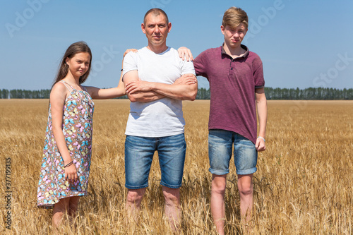 Pre-teen son and daughter with their single father, kids lay hands on his shoulders, looking at camera, full length portrait at wheat field