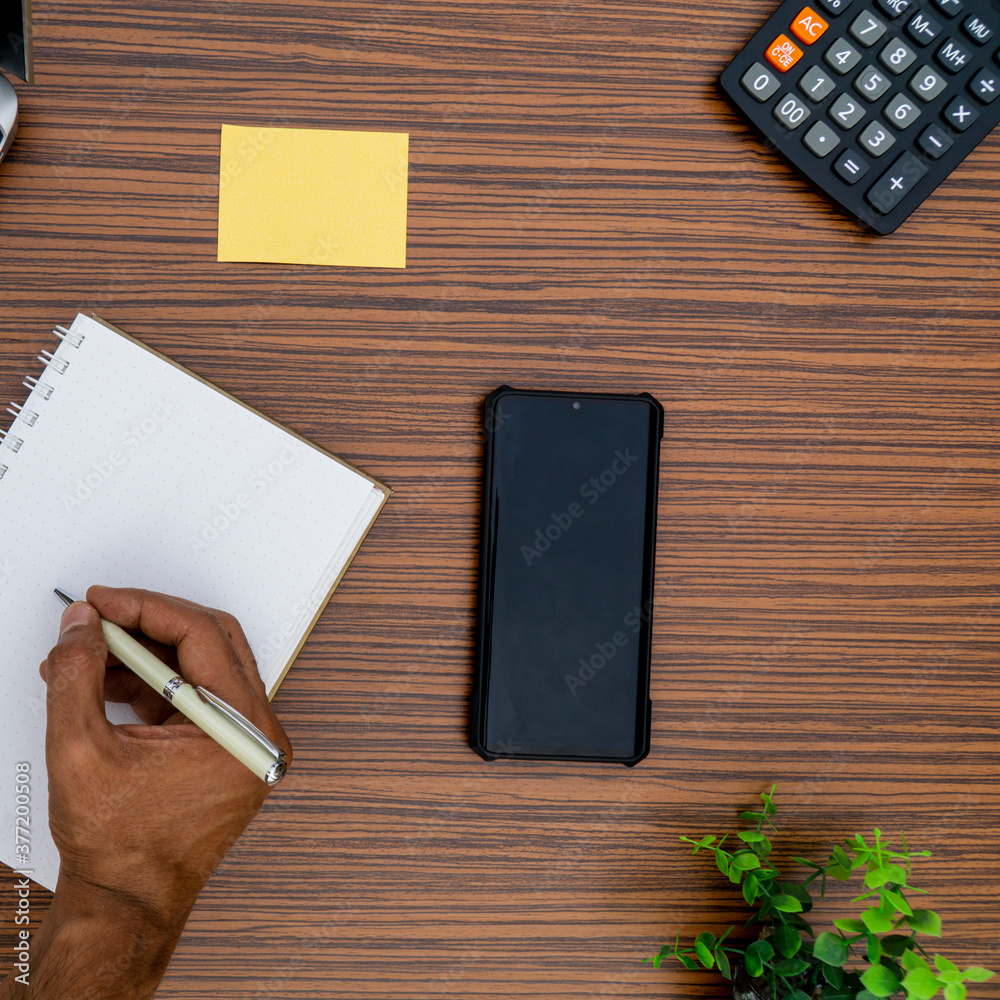 Writing on a notepad while working from home. A lap top, a mobile, calculator and plant are also on display on this brown striped working table. Writing on a notepad while working from home. A lap top, a mobile, calculator and plant are also on display on this brown striped working table.