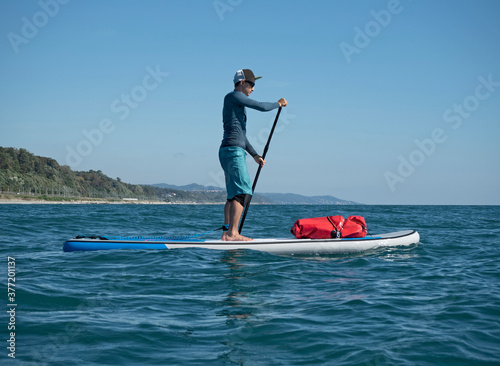 Active man rowing on SUP touring with waterproof bag at sunny day