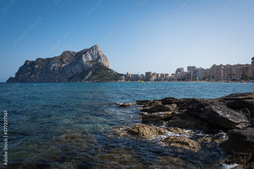 Naklejka premium View from the rocks of the coastline with the urban beach of Calpe and the crag in the background, Alicante, Spain