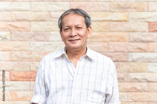 Portrait of smiling asian senior man standing in front of brick wall background with self confident manner. Closeup face of elder smile with gray hair.