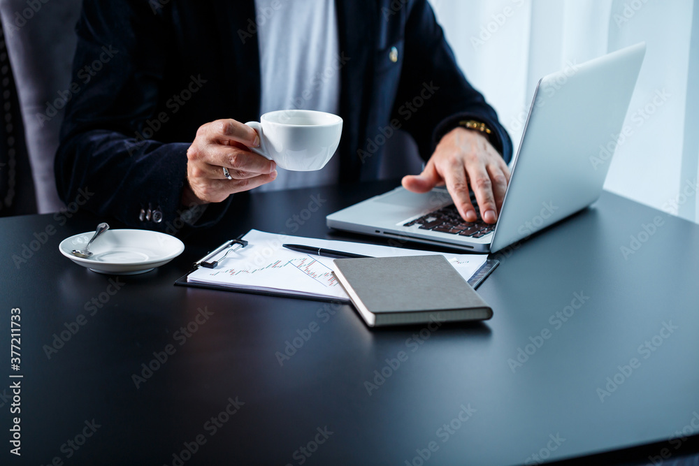 An adult male businessman is working on a new project and looking at the stock growth charts. Sits at the table by the large window. Looks at the laptop screen and drinks coffee. An adult male businessman is working on a new project and looking at the stock growth charts. Sits at the table by the large window. Looks at the laptop screen and drinks coffee.