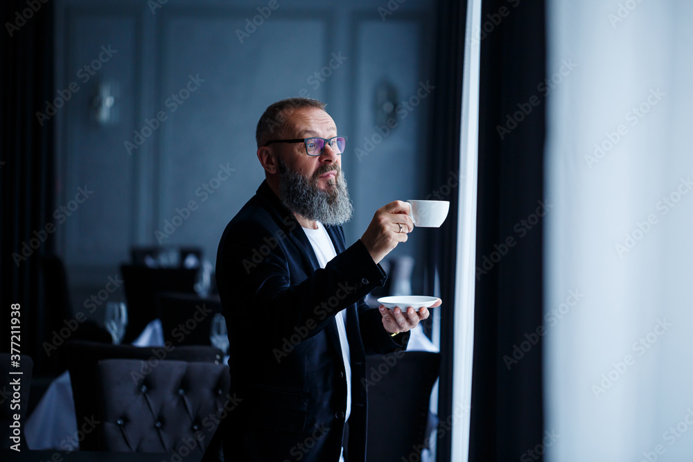 An adult male mentor, director, businessman in glasses and a suit drinks coffee and has a rest. Working day concept An adult male mentor, director, businessman in glasses and a suit drinks coffee and has a rest. Working day concept