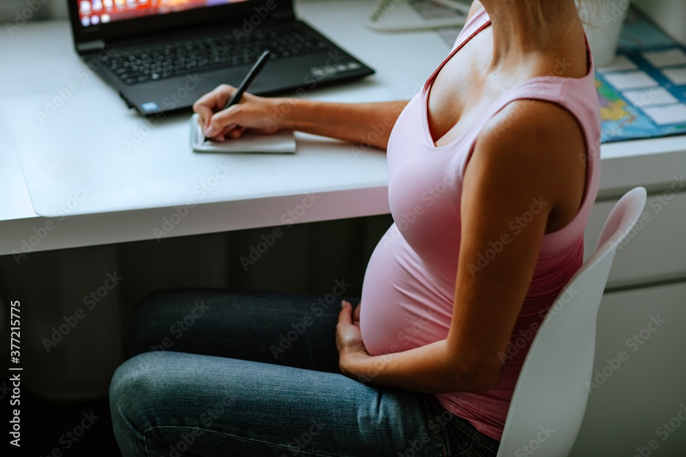 Pregnant woman sitting at the white table and writing down to the notebook.