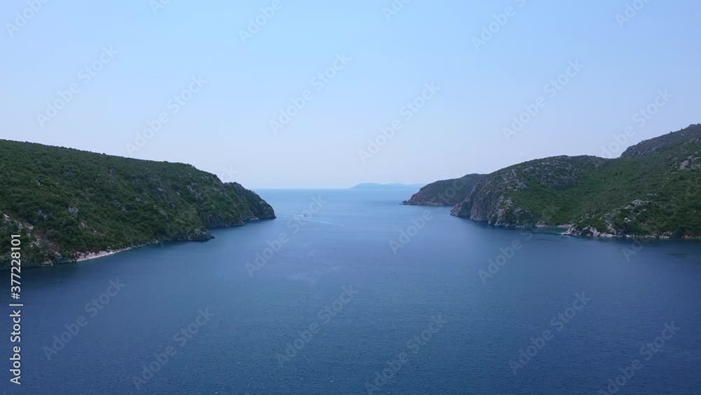 Aerial view of bay with passage between to hills covered by pine trees and open blue sea behind.