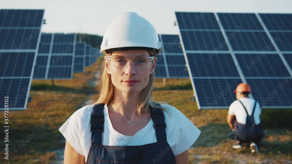 Portrait of nice-looking cheerful female blonde engineer in uniform standing by solar batteries. Woman worker cooperating with colleague on ecological solar farm. Portrait of nice-looking cheerful female blonde engineer in uniform standing by solar batteries. Woman worker cooperating with colleague on ecological solar farm.