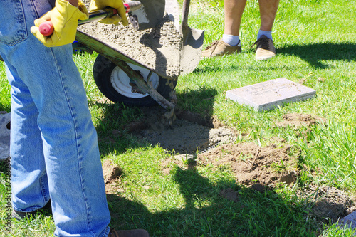 People are pouring wet concrete from a wheelbarrow with the help of a shovel  for the base of a gravestone