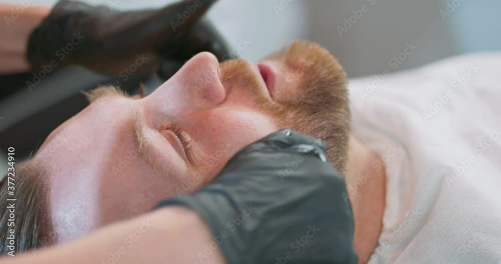 Closeup young bearded man's head, on the barber's chair, with cape, and female barber's hands in black gloves applies oil and cosmetics to the client's beard. Closeup young bearded man's head, on the barber's chair, with cape, and female barber's hands in black gloves applies oil and cosmetics to the client's beard.