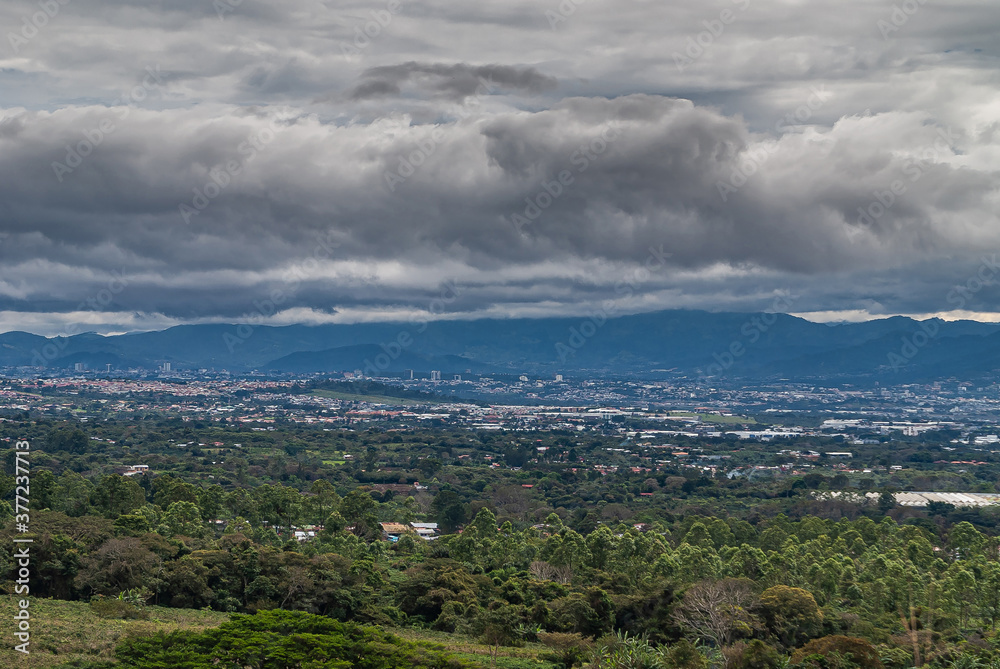 Alajuela Province, Costa Rica - November 28, 2008: San Jose City skyline in its valley under heavy thick cloudscape, seen from up the mountains. Green foliage in front.