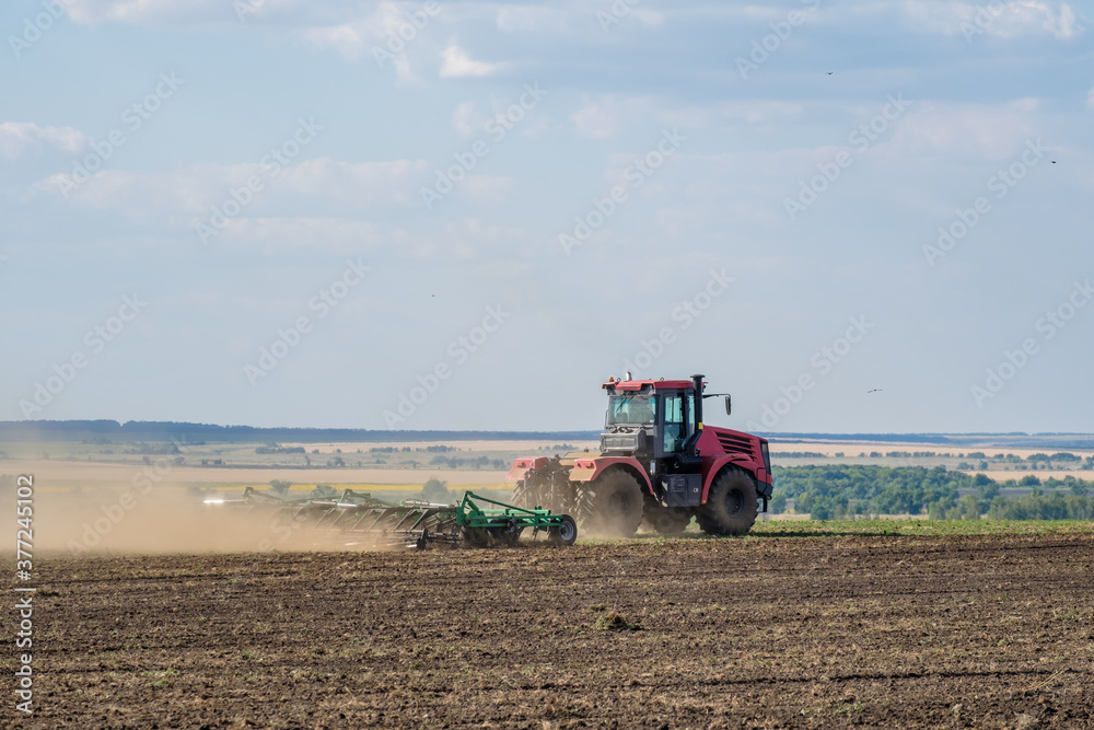 A red farm tractor in a cloud of dust cultivates the soil in the field with a cultivator after harvest. Summer sunny day. Fertile land. Modern agricultural machinery. Copy space. High quality photo A red farm tractor in a cloud of dust cultivates the soil in the field with a cultivator after harvest. Summer sunny day. Fertile land. Modern agricultural machinery. Copy space. High quality photo
