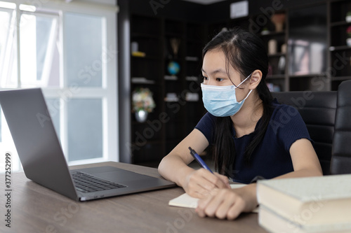 Asian student wearing protective mask,taking notes from laptop computer and textbooks in library at school,writing do homework,concentrated child girl make notes in the book prepare for test or exam