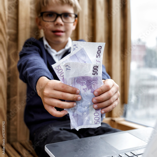 the boy sits at the computer and shows money to the camera. online shopping by children