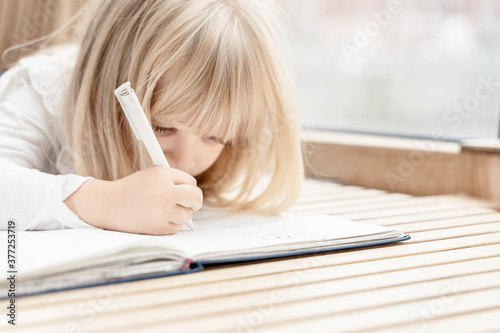 beautiful little blonde girl lies on the floor and writes something in a notebook. low contrast image with copy space. selective focus on hand
