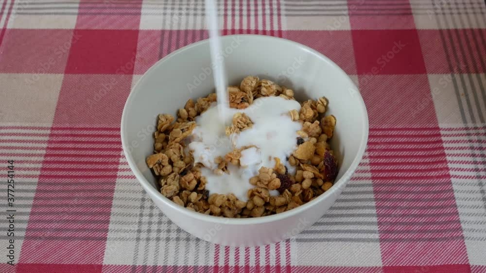 Pouring yogurt in bowl of whole grain muesli on a table with a red and white tablecloth. Pouring yogurt in bowl of whole grain muesli on a table with a red and white tablecloth.